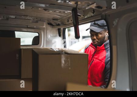 POV inside delivery van. Tired focused Black male courier in red jacket, black hat, and wireless headphones choosing the right parcel to deliver. High quality photo Stock Photo