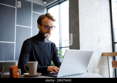 A young red-haired guy, a programmer or entrepreneur in glasses, works in a stylish cafe behind a laptop. Freelancer works remotely. Online Stock Photo