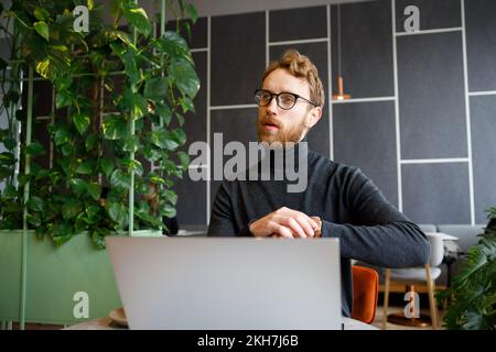 A young red-haired guy, a programmer or entrepreneur in glasses, works in a stylish cafe behind a laptop. Freelancer works remotely. Online Stock Photo