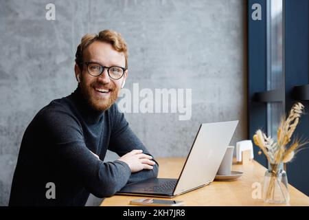 A young red-haired guy, a programmer or an entrepreneur in glasses, in a stylish cafe behind a laptop, looks at the camera and smiles. Freelancer Stock Photo