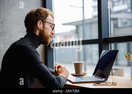 A young red-haired guy, a programmer or businessman in glasses, is sitting in a stylish cafe at a laptop and is talking through a webcam. Freelancer Stock Photo