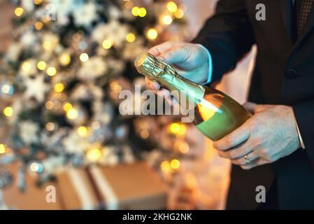 Close up shot of a man in suit opening a bottle of champagne, there is a christmas tree on the background. Stock Photo