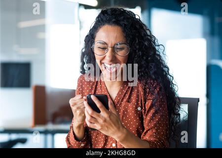 Happy and joyful hispanic business woman working inside office reading online message on phone, pleased and shocked celebrating victory success, close up photo of office worker in glasses. Stock Photo