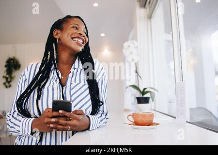Young businesswoman smiling and looking away while holding a smartphone in a coffee shop. Happy young black businesswoman enjoying her coffee break in Stock Photo