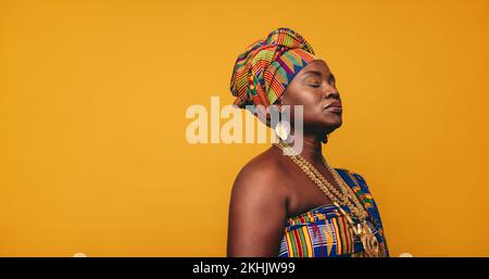 Ghanaian woman wearing traditional clothing against a yellow background. Mature black woman dressed in colourful Kente cloth and golden jewellery. Con Stock Photo