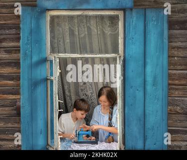 Portrait of girl and boy kids tailor sew making doll's clothes on a children's sewing machine in the window of an old wooden house. Hobby, art and chi Stock Photo
