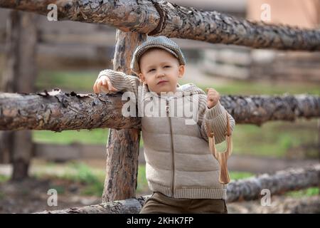 Bully boy kid with a slingshot aims at someone near a fence in the village outdoors. Rustic barefoot child boy with a hat shoots a slingshot. Stock Photo