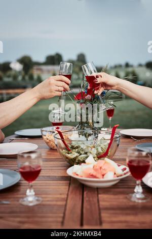 Couple making toast during summer outdoor dinner in a home garden. Hands holding wine glasses with red wine over table with dishes Stock Photo