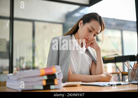 Beautiful stressed millennial Asian businesswoman or female boss sitting at her desk, working on her paperwork, feeling upset and dissatisfied with he Stock Photo