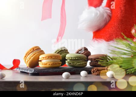 French macaroons of various flavors on slate plate on a wooden table and Christmas hat with white isolated background. Front view. Stock Photo