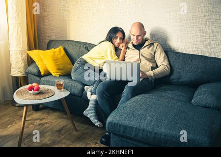 Multi ethnic couple in love spends leisure time with a laptop at home. Multiracial girlfriend and caucasian boyfriend look at the laptop screen sittin Stock Photo
