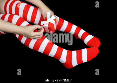 Girl in Christmas knee socks sitting with red gift box on black background. Female outfit for New Year celebration Stock Photo
