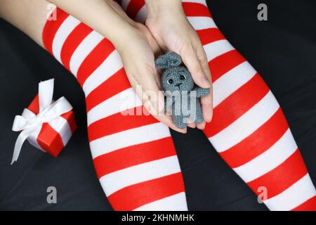 Girl in Christmas knee socks sitting with grey knitted rabbit in hands. Symbol of Chinese New Year 2023, female outfit for celebration Stock Photo