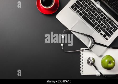 Laptop on a black desktop with a cup of coffee and a green apple and a stethoscope for a doctor, working on social networks, writing in a notebook. Vi Stock Photo