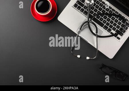 A laptop on a black work desk with a cup of coffee and black glasses on the table and a stethoscope for a doctor working on social networks. View from Stock Photo