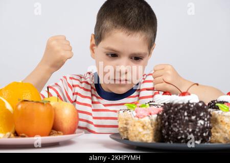 5 year old cute boy sits in front of fruits and cakes and chooses what to eat Stock Photo