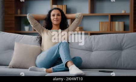 Calm happy in good mood caucasian hispanic latino 30s woman resting on couch new sofa in living room stretching hands behind head healthy lady Stock Photo