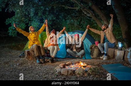 Happy friends celebrate and have fun laughing together with arms raised bonfire on camping Stock Photo