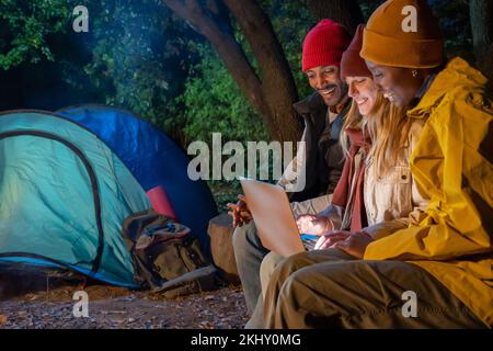 Smiling friends looking at a laptop computer while camping in a tent in the forest at night. Stock Photo