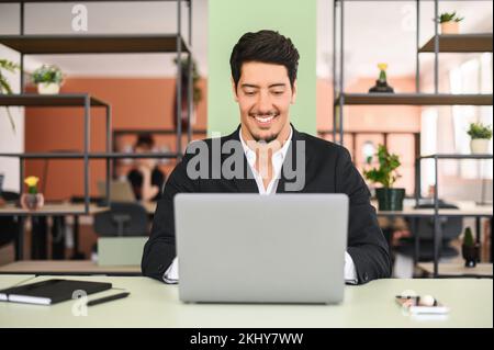 Positive latin dark haired businessman wearing formal suit sits at the desk in contemporary office space, using laptop, conducts business correspondence, replying emails. Hispanic man office employee Stock Photo