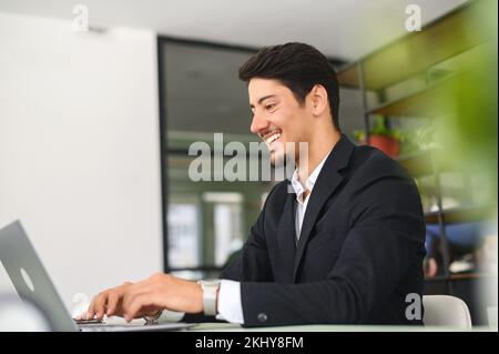 Happy smart young hispanic businessman wearing formal suit using the laptop computer at modern office. Smiling latin male office employee at the workplace is typing Stock Photo