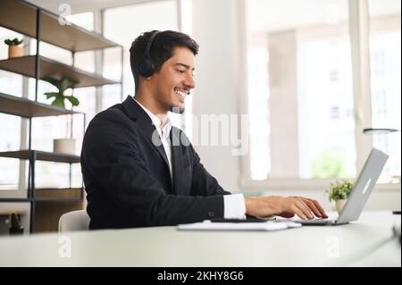 Young man wearing wireless headset using a laptop computer for communication with customers or colleagues sitting at the desk in contemporary office. Maale support worker talks online. Side view Stock Photo