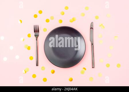 Stylish table setting with black plate, cutlery on pink background with golden confetti. Christmas, birthday, holiday concept. Top view, flat lay. Stock Photo
