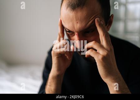 Close-up face of exhausted male sitting alone at home touching head, feeling sad, frustrated, depressed, suffering from headache after quarrel with Stock Photo