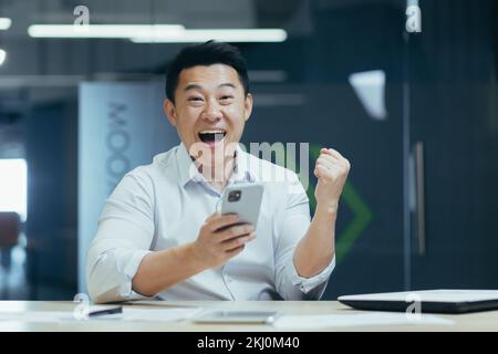 A young man, an Asian office worker, plays online games on the phone, rejoices at the victory. He points with his hand, yes, the winner. Sitting in the office at the desk, looking at the camera. Stock Photo