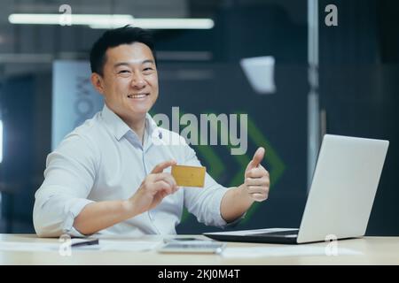Portrait of a young Asian businessman. Posing, smiling at the camera, holding a credit card and pointing super, sitting in the office at the desk Stock Photo