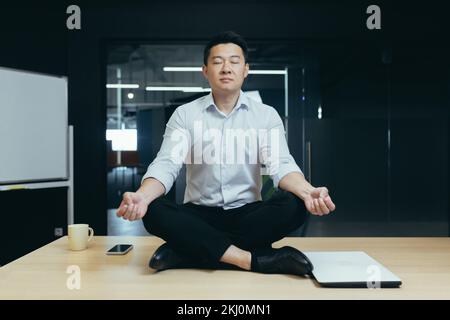 Rest break at the workplace. A young Asian man sits in the office on the desk in the lotus yoga position with closed eyes, meditates, relaxes during working hours. Stock Photo