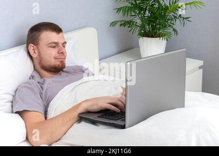 A man with laptop is lying in bed. Freelancer works remotely from home. Distance education. Online shopping without leaving home. Internet surfing. Gambling addiction. Black Friday sale. Cyber Monday. Stock Photo