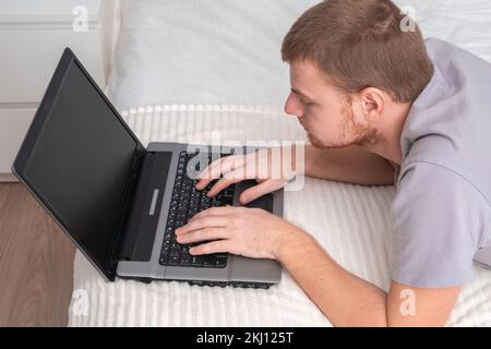 A man with laptop is lying in bed. Freelancer works remotely from home. Distance education. Online shopping without leaving home. Internet surfing. Gambling addiction. Black Friday sale. Cyber Monday. Stock Photo