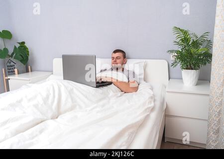 A man with laptop is lying in bed. Freelancer works remotely from home. Distance education. Online shopping without leaving home. Internet surfing. Gambling addiction. Black Friday sale. Cyber Monday. Stock Photo