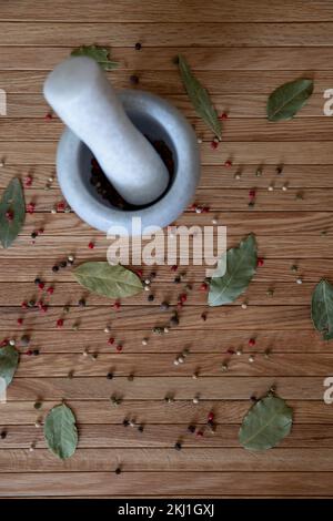 A stone mortar for spices stands on a wooden table. Scattered on the table are lavender leaves and a mixture of peppercorns. Stock Photo