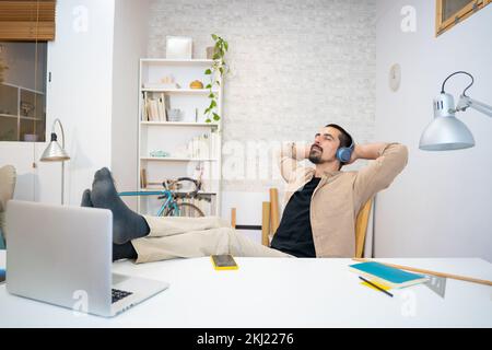 Positive young man keeping hands behind head while listening to music in headphones at home Stock Photo