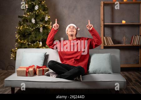Handsome young man shows the forefingers up on copy space sitting at Christmas home. Happy student at Santa hat Stock Photo
