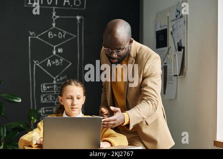 African American teacher talking to schoolgirl while she using laptop at desk at lesson Stock Photo