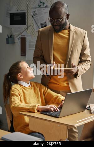 Smiling schoolgirl talking to teacher while using laptop at desk during IT technology lesson Stock Photo