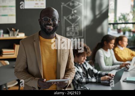 Portrait of African American IT teacher smiling at camera standing in classroom with students using computers in background Stock Photo