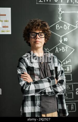 Portrait of schoolboy with curly hair and eyeglasses looking at camera while standing with his arms crossed against blackboard at class Stock Photo