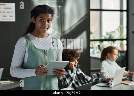 Portrait of African American schoolgirl with tablet pc looking at camera standing at classroom with her classmates in background Stock Photo