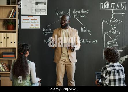 African American teacher standing near the blackboard with charts and explaining material to children at IT lesson Stock Photo