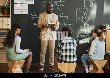 African American teacher using tablet pc and giving lecture to children standing against blackboard at IT lesson Stock Photo
