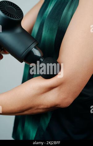 a young caucasian man, in sportwear, uses a massage gun to massage the muscles of his arm, near to his elbow Stock Photo