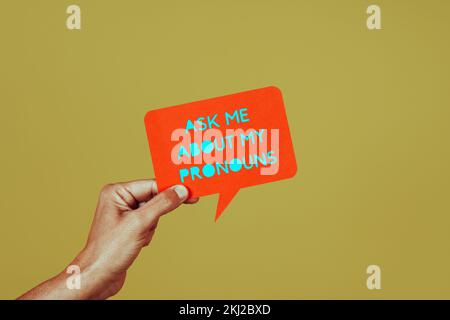 closeup of a person holding an orange paper sign in the shape of a speech bubble with the text ask me about my pronouns, on a green background Stock Photo