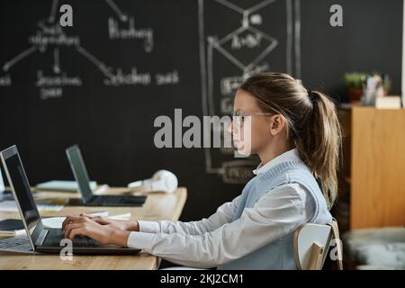 Schoolgirl in eyeglasses concentrating on her online work, she typing on laptop and learning to use new software at class Stock Photo