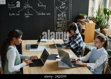 Group of school children working with interface on computers at table in classroom with big blackboard with charts Stock Photo