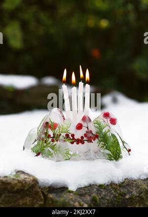 Handmade frozen Christmas wreath in shape of bundt cake made of ice,  red rose hips berries, holly berry branches and thuja twigs with four burning ca Stock Photo