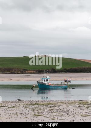 A small old fishing boat is anchored, shallow water at low tide on a cloudy day. Hills under a cloudy sky. Silt and seaweed. Landscape. Stock Photo